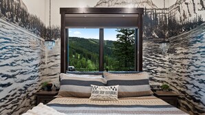 Een slaapkamer met een groot raam, een bed met beddengoed met een patroon, een kussen met de tekst 'NEVER STOP EXPLORING', en een bergmuur op de wanden.