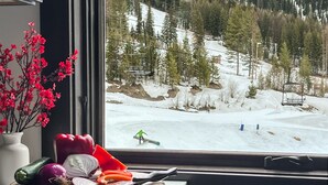 Uitzicht vanuit een raam op een besneeuwd berglandschap, met een skilift, een skiënde persoon en een tafel met groenten en een mes.