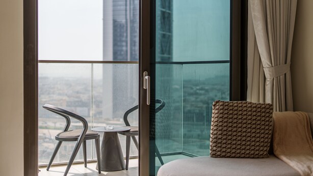 Een balkon met een glazen deur, twee stoelen, een klein tafeltje en een gekleurd kussen op een bed.