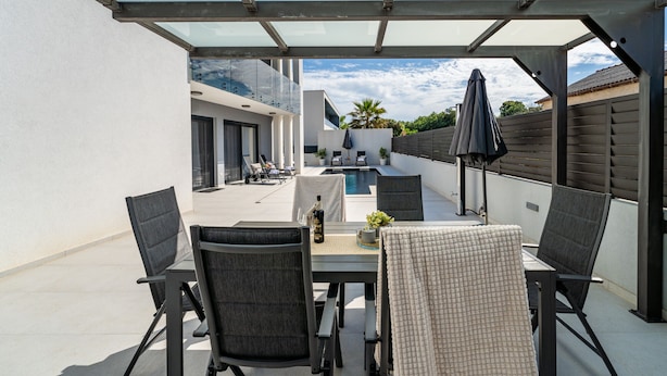 Een overdekt terras buiten met een zwembad, een eettafel en stoelen.
