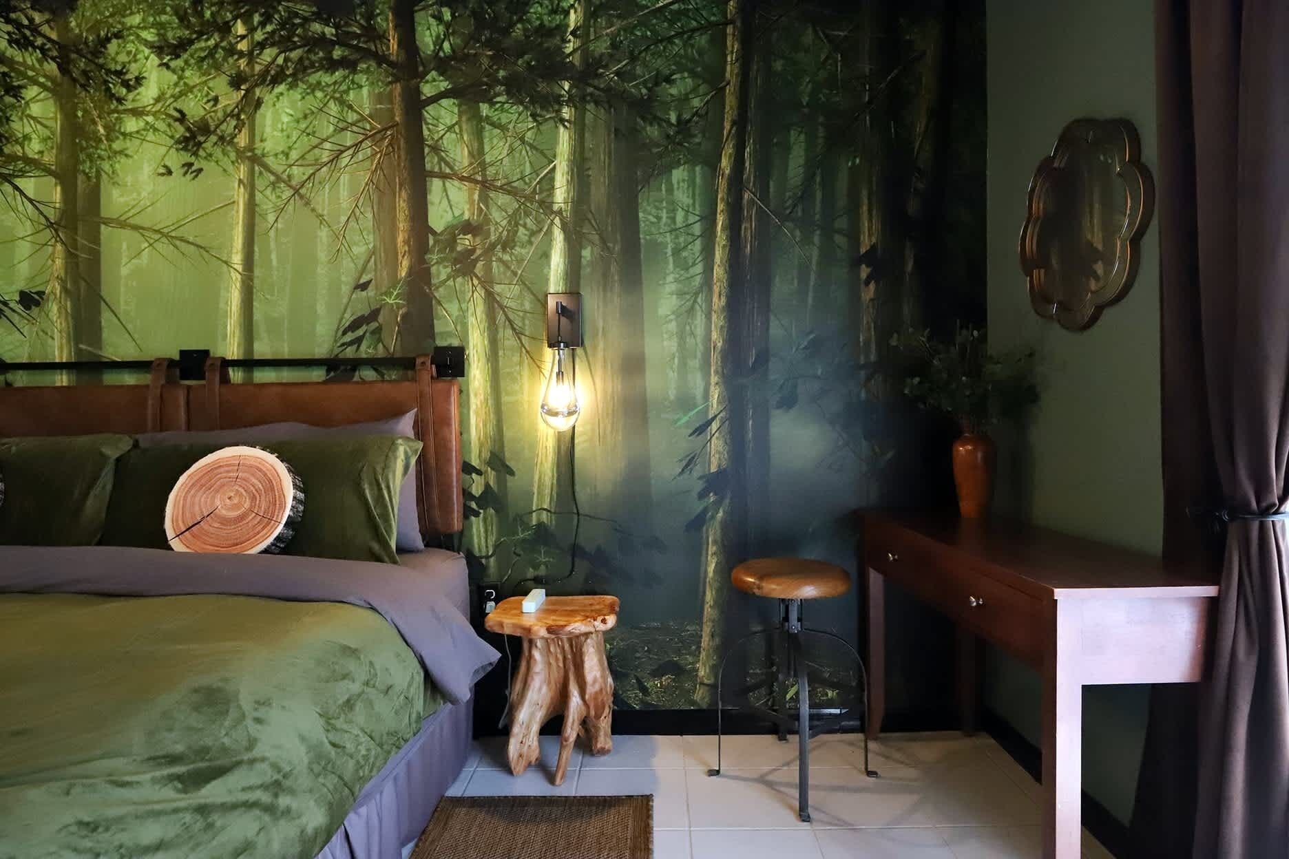 Une chambre à coucher comprenant un lit, une table de chevet, un tabouret, une lampe fixée au mur et une grande fresque murale sur le thème de la forêt.
