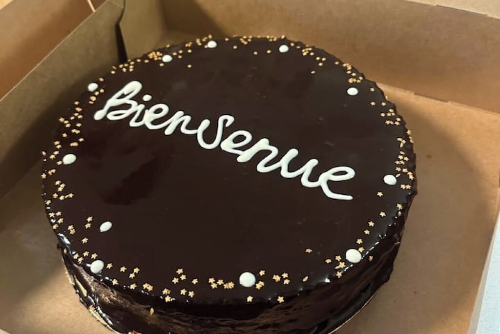 A round chocolate cake with "Bienvenue" written on top, decorated with small white and gold edible decorations.