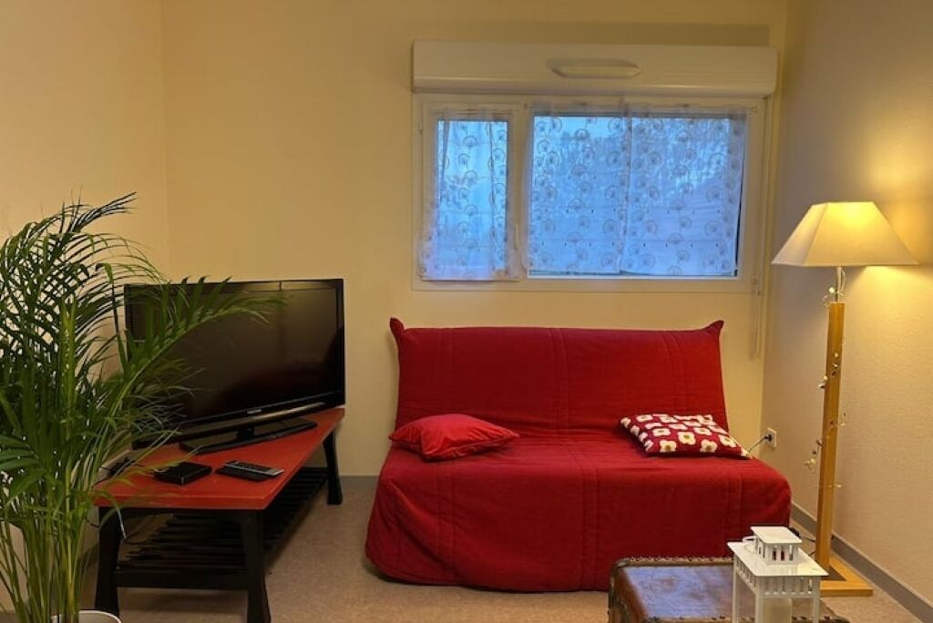 A room with a red sofa, a television on a stand, a small table, a lamp, and a window with curtains.
