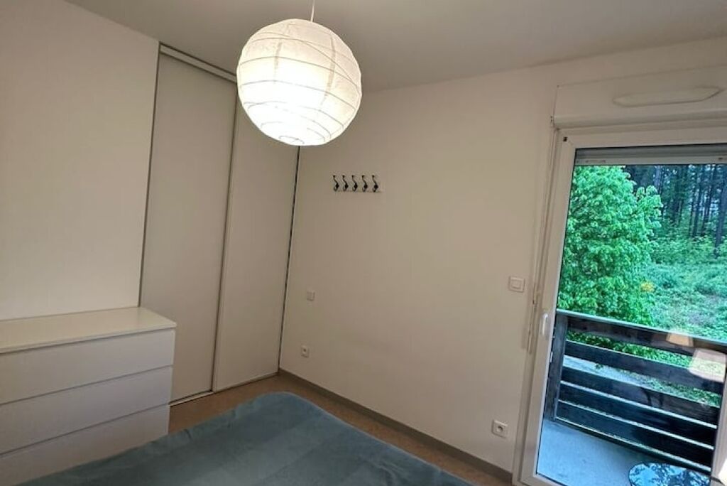 Ein Schlafzimmer mit Bett, Kleiderschrank, Pendelleuchte, einem Fenster mit Blick ins Grüne und einer Tür.