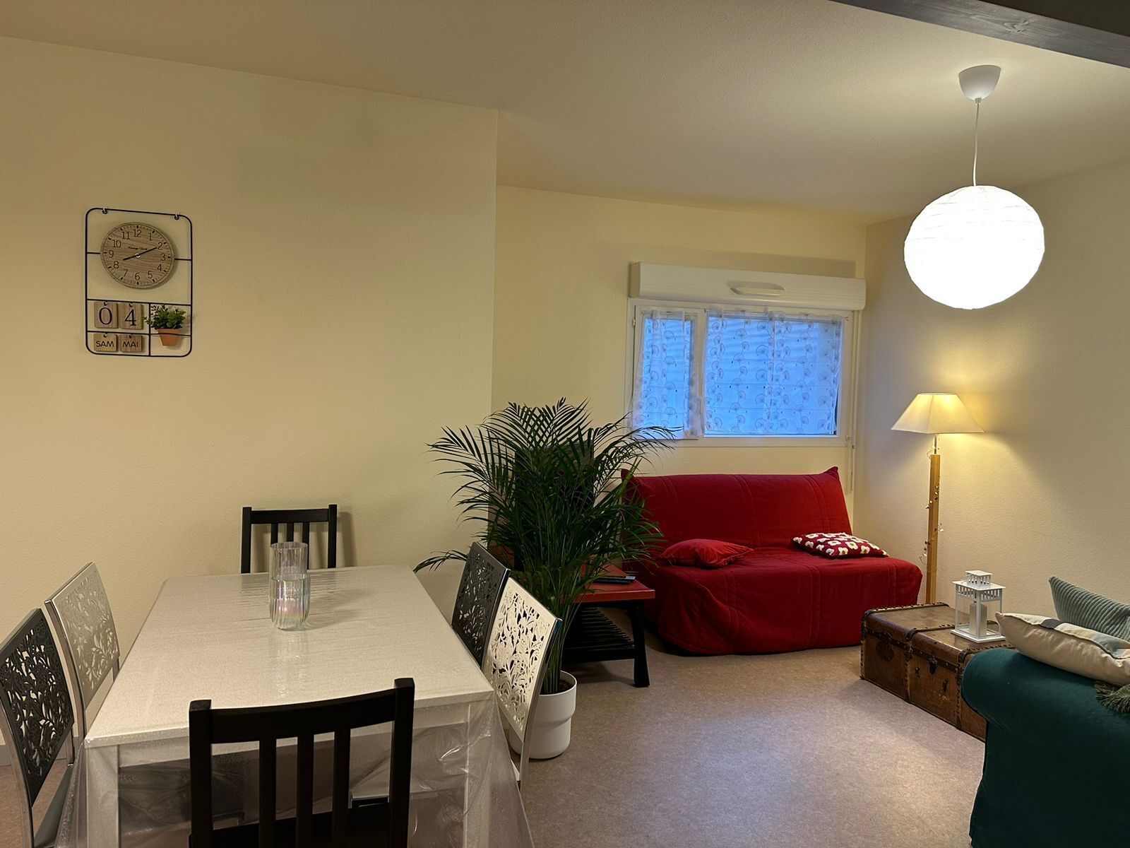 A dining area with a table, chairs, and a clock on the wall. There is a window with curtains and a lamp on a side table.