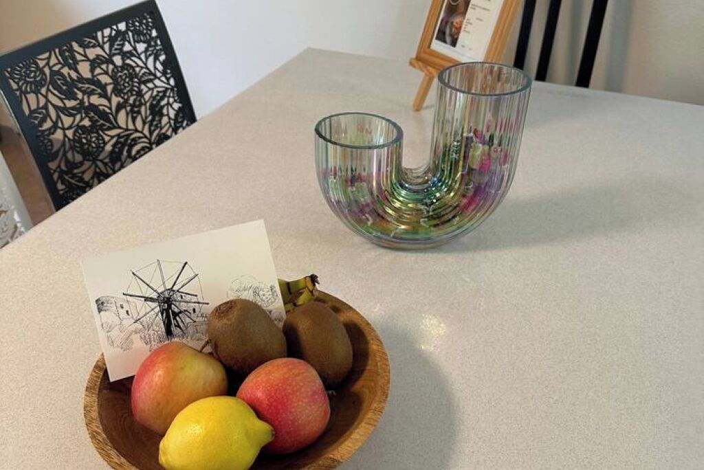 A wooden bowl with apples and a lemon on a table, a glass vase with flowers, and a framed picture.