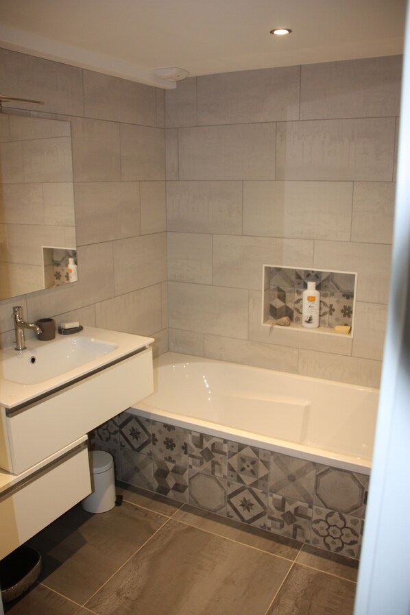 A modern bathroom with a white sink, a bathtub, and a shower area. There are decorative tiles on the walls and a small shelf with toiletries. - A spacious, charming family chalet surrounded by nature. (Damprichard)