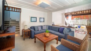 A modern living room with a sofa, coffee table, and wicker chairs. There is a TV on a stand, a small table with a lamp, and framed pictures on the wall. - Bz92 Casa 600m Rua das Pedras 4 suítes (Buzios)