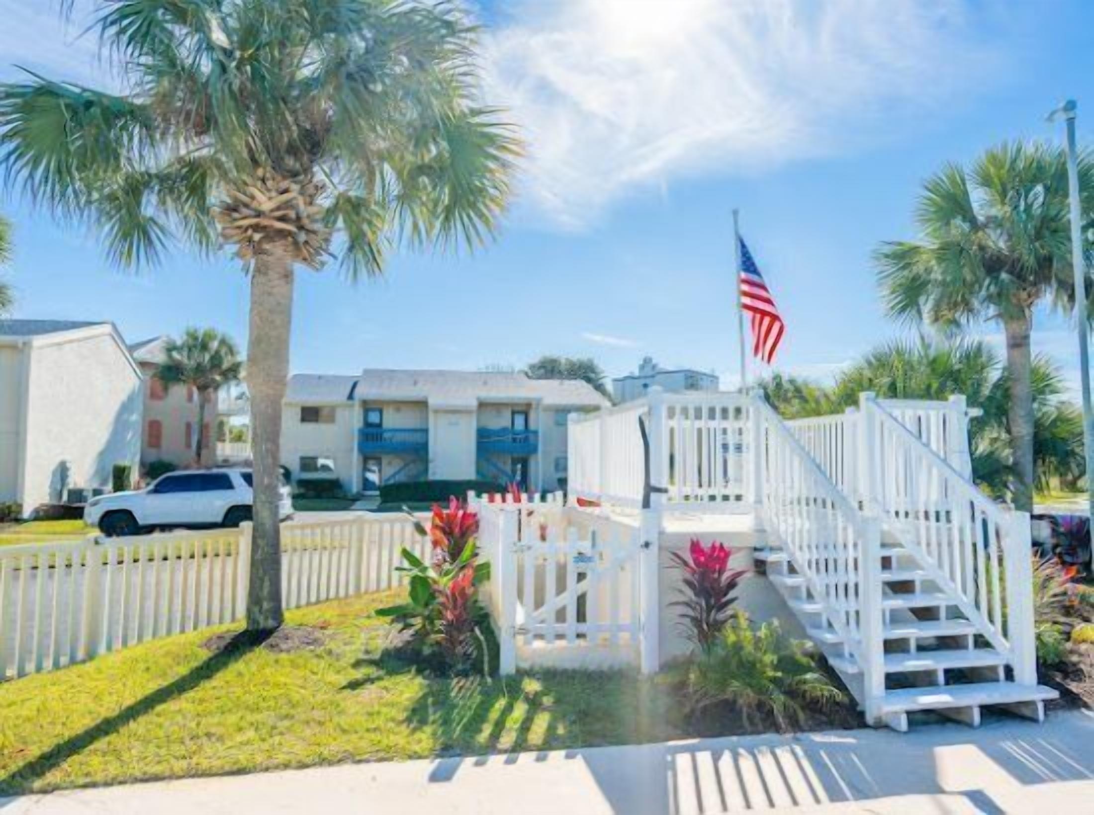 A hotel entrance with a white fence, palm trees, and an American flag.