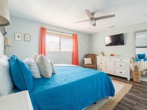 A bedroom with a bed, a ceiling fan, a window with curtains, a dresser, and a television on the wall.