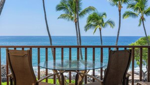 A balcony with two chairs and a glass table overlooking the ocean, with palm trees and a clear sky in the background. - Summer Deal Oceanfront Kona 1BR Condo with Pool and Beach Access (Kailua-Kona)