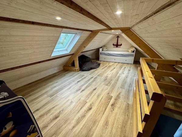 A loft bedroom with a sloped ceiling, wooden flooring, a bed, a desk, and a window.