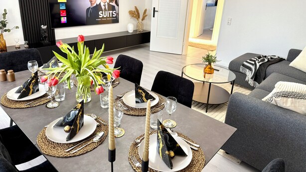 Una sala de estar moderna con un televisor de pantalla plana mostrando una serie de Netflix, un área de comedor con una mesa, sillas y un jarrón de tulipanes.