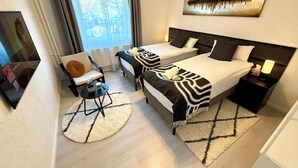 Habitación de hotel con dos camas, una silla, una mesita, una lámpara y un cuadro en la pared.