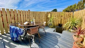 A wooden deck with a round table and wicker chairs, surrounded by potted plants and a wooden fence. - Padstow Holiday Home 2 Bedrooms, wifi, rear decked patio, pets welcome - parking (Padstow)