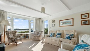 A spacious living room with a large window, a sofa, a patterned armchair, and a bookshelf.