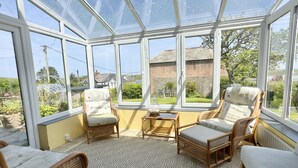 Un invernadero con muebles de mimbre y suelo estampado, con ventanas grandes que ofrecen vistas a un jardín y edificios.