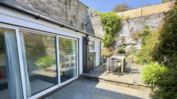A patio area with a stone floor, a table with chairs, and a view of a garden with a stone wall and greenery. - 36 High Street -  a two storey property that sleeps 6 guests  in 3 bedrooms (Padstow)