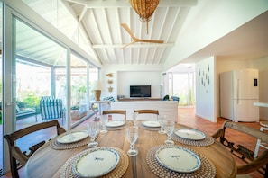 Comedor con una gran mesa de madera dispuesta para seis personas, rodeada de sillas. Se observa un ventilador de techo y una lámpara de fibra trenzada arriba.