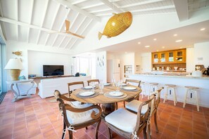 A spacious dining area with a round table set for six, surrounded by wooden chairs with cushioned seats. A large woven fish hanging light fixture is visible above the table. - Bz93 Cond pé na areia piscina privativa (Buzios)