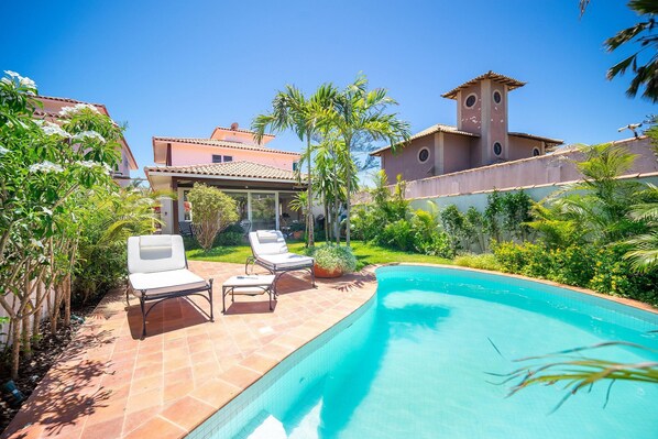 A swimming pool with lounge chairs, surrounded by lush greenery and a large house with a tower. - Bz93 Cond pé na areia piscina privativa (Buzios)