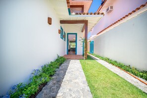 A narrow pathway leading to a building entrance with a tiled roof and a small garden area with grass and plants. - Bz93 Cond pé na areia piscina privativa (Buzios)
