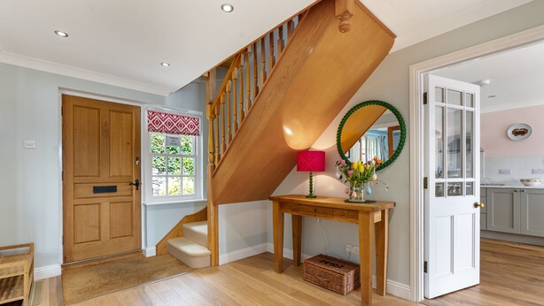 A wooden staircase leading to a second floor, a wooden console table with a vase, a door with a wreath, and a kitchen area visible through an open door.