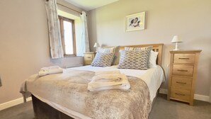 A neatly made bed with a beige blanket and two pillows, a wooden nightstand with a lamp, a dresser with drawers, and a framed picture on the wall.