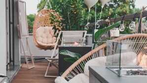 Un terrazzo in legno con mobili in vimini, tra cui una poltrona sospesa e una poltrona imbottita, e un tavolo in vetro con un elemento decorativo a fuoco.