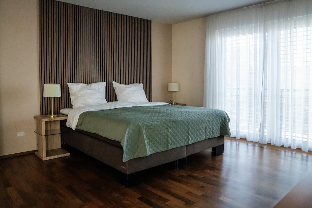 A modern hotel room with a large bed, wooden flooring, a striped wall panel, and a large window with sheer curtains.