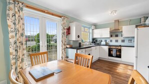 A kitchen with white cabinets, a wooden table, and a window with curtains. - Kegyn - Harlyn Bay Cornwall,  sleeps 5 guests & accepts pets (Harlyn Bay)
