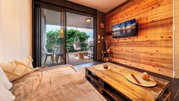 Een moderne kamer met een bed, een houten tafel met een kom en een hoed, een aan een houten wand bevestigde televisie, en een schuifdeur die leidt naar een buitenruimte met een tafel en stoelen.