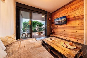A modern room with a bed, a wooden table with a bowl and a hat, a TV mounted on a wooden wall, and a sliding glass door leading to an outdoor area with a table and chairs. - L'Ecrin Bohème (Saint-Paul)