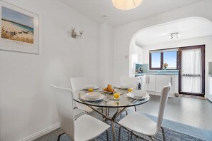 Ein moderner Essbereich mit einem Glastisch, weißen Stühlen und Frühstücksangeboten. An der Wand hängt ein gerahmtes Bild von einem Strand.
