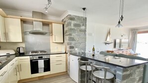 A modern kitchen with a central island, bar stools, and a stone accent wall. - 14 The Old Boatyard -with parking and  Incredible Estuary Views (Padstow)