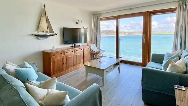 A living room with a TV, wooden cabinets, and a view of the sea. - 14 The Old Boatyard -with parking and  Incredible Estuary Views (Padstow)