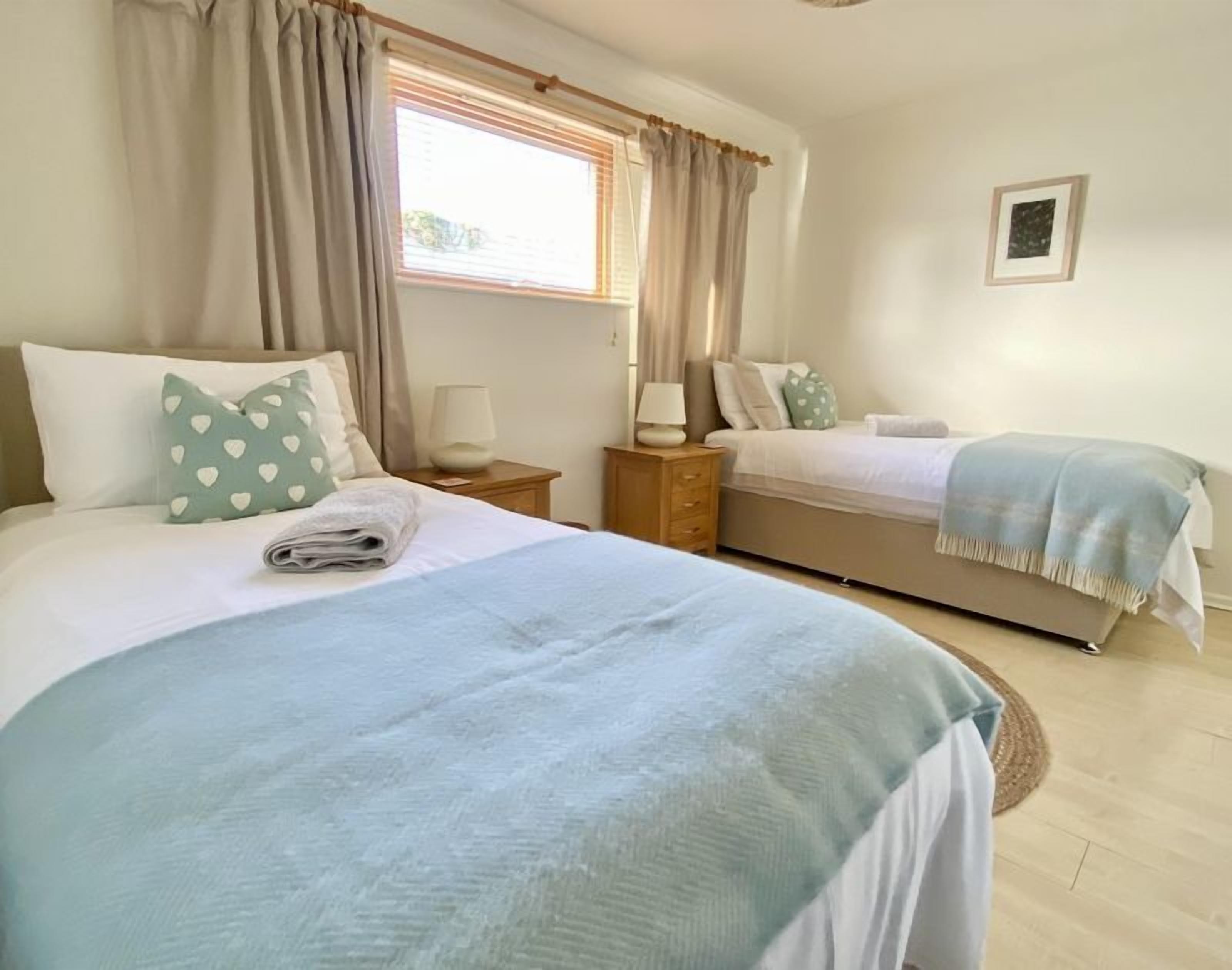 A bedroom with two beds, a window with curtains, a bedside table with a lamp, and a framed picture on the wall.