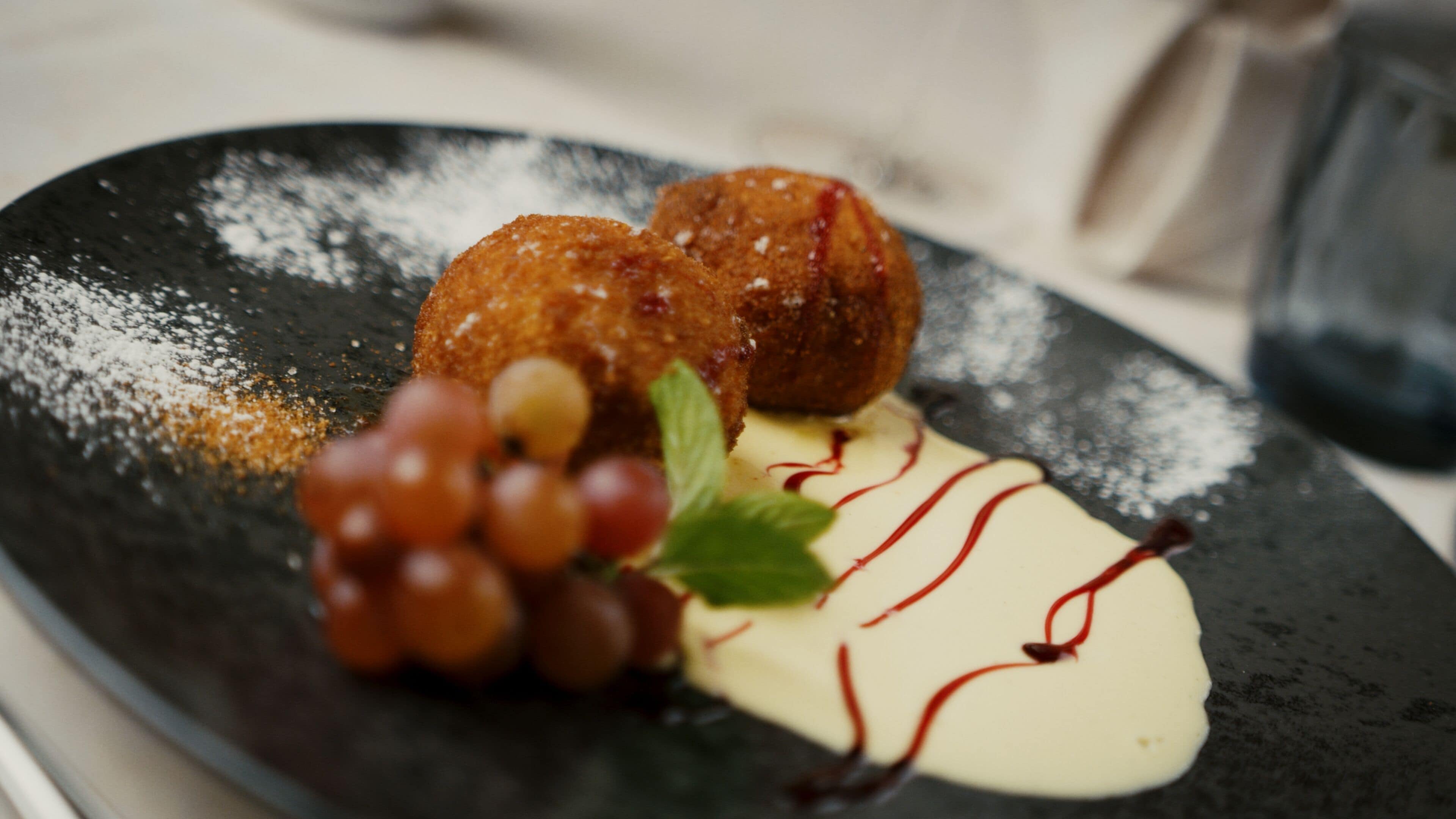 A dessert plate with a dark plate, a glazed pastry, grapes, and a mint leaf.