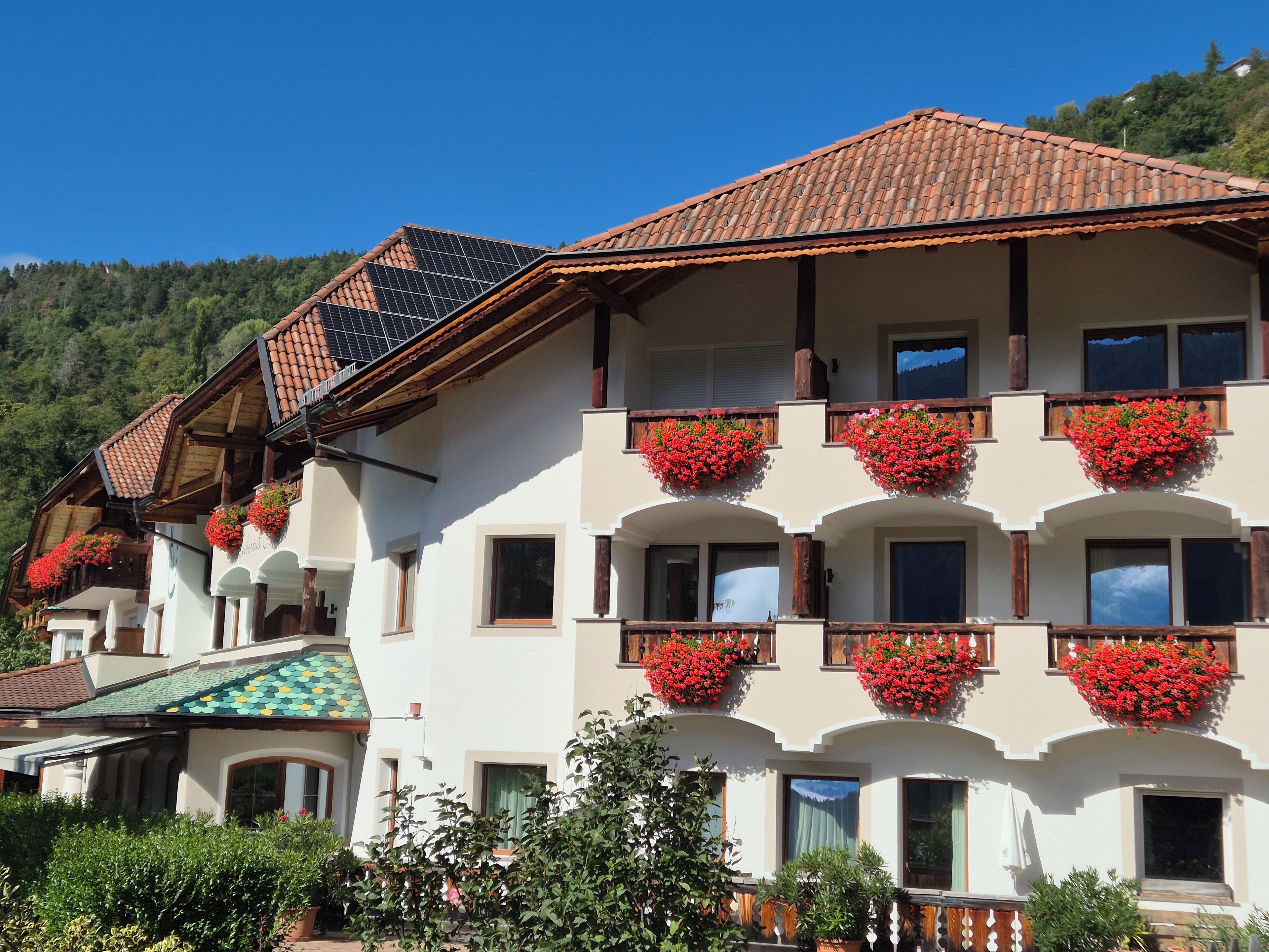 Un edificio de varios pisos con balcones decorados con flores rojas, techo de tejas y montañas al fondo.