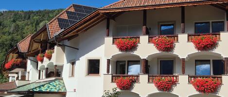A multi-story building with balconies adorned with red flowers, a tiled roof, and a mountainous backdrop.