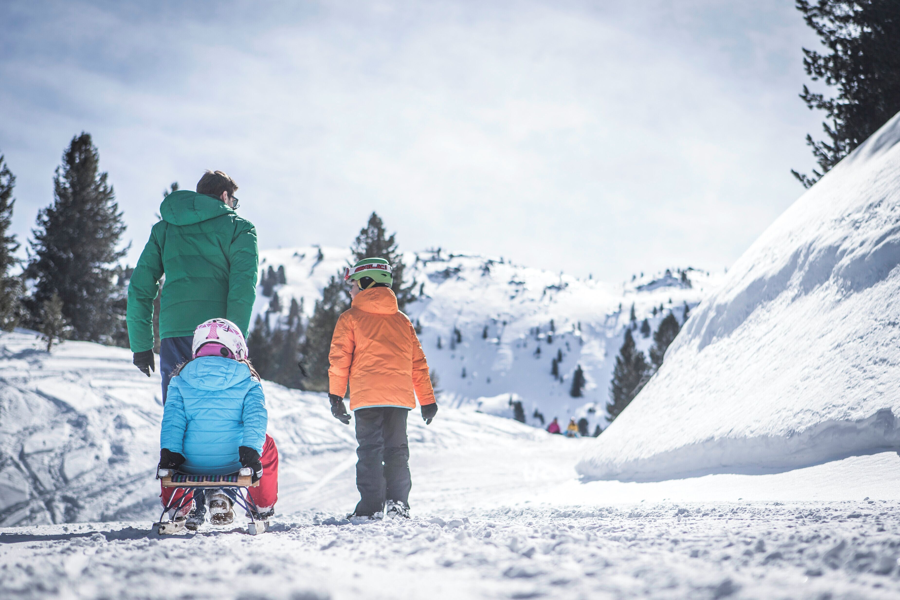 Escena nevada de una montaña con una persona con chaqueta verde y un niño con chaqueta naranja caminando por un sendero cubierto de nieve.