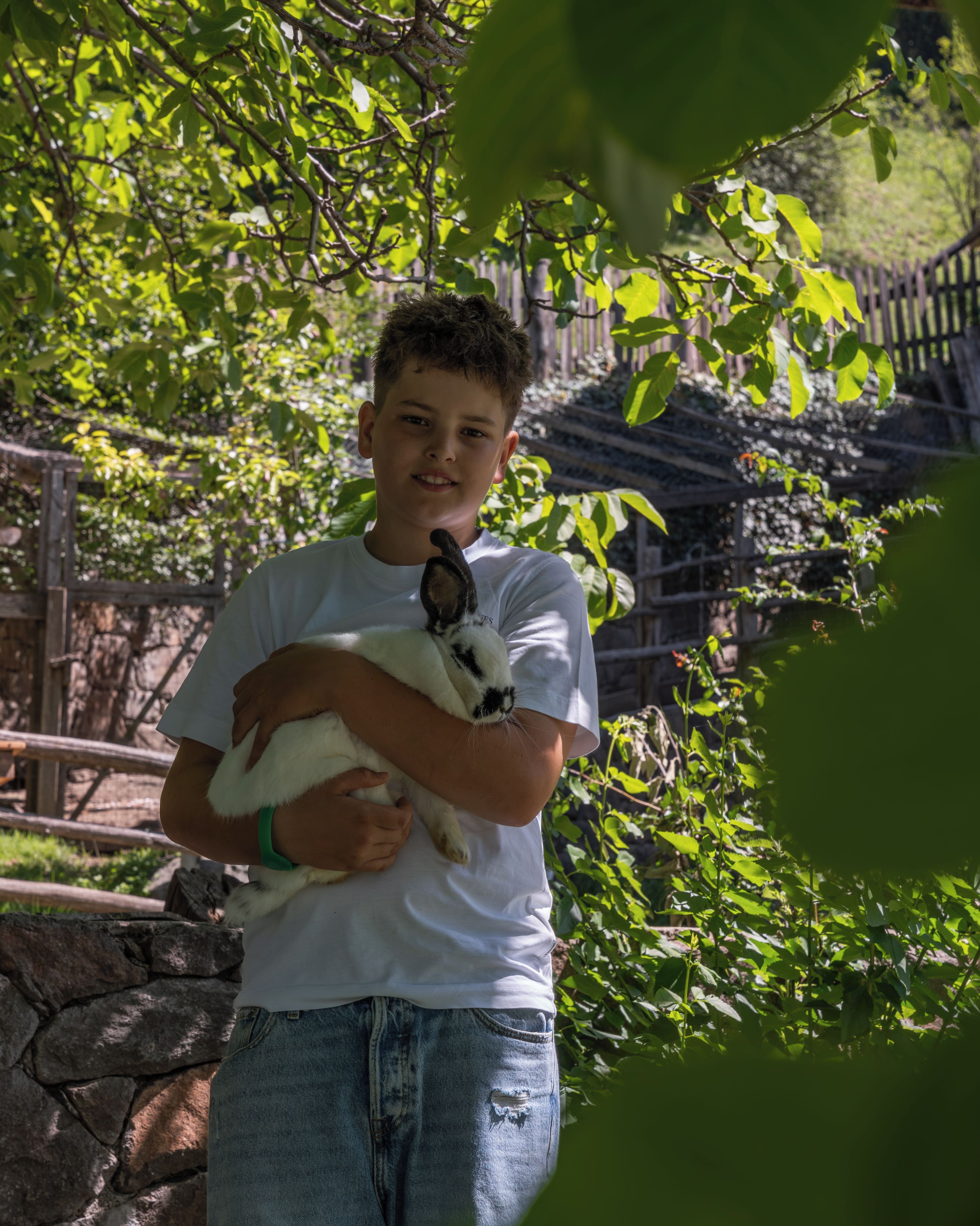 A person holding a white and black rabbit in a natural setting with greenery and a wooden fence.