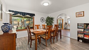 Un comedor con una mesa y sillas de madera, un ventanal con cortinas, una planta en maceta y una estantería llena de libros.