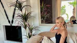Una mujer sentada en un sofá color beige en una sala de estar moderna con piso de mármol, un televisor de pantalla plana montado en la pared y varias plantas en macetas.