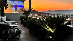 Un balcón con vista a una puesta de sol sobre un paisaje urbano, con mobiliario exterior y plantas en macetas.
