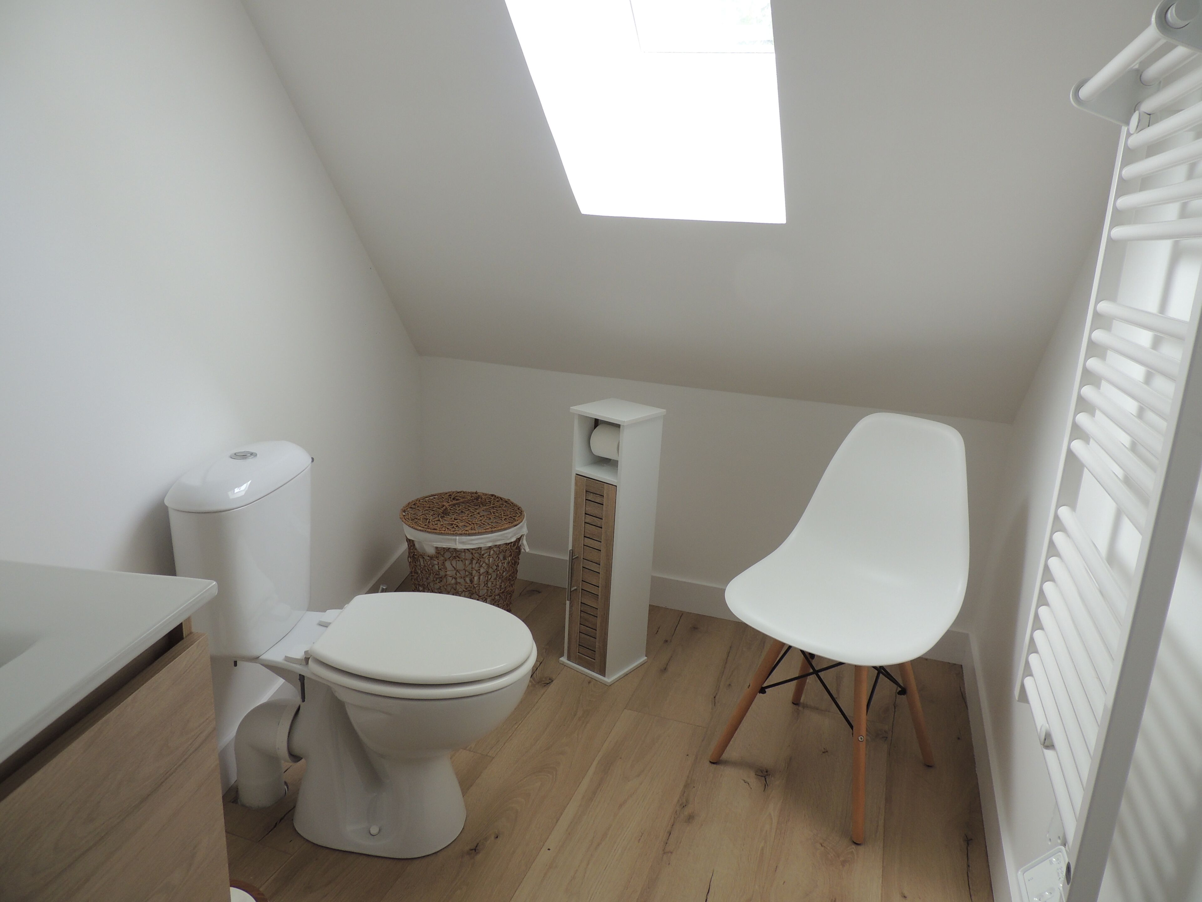 A small, modern bathroom with a toilet, a wicker basket, a white chair, and a skylight.