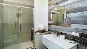 A modern bathroom with a glass shower enclosure, a white vanity with a round mirror, and a wooden wall panel. - Camera ampia matrimoniale/tripla (Vitorchiano)