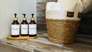 A bathroom with a wooden shelf holding three bottles of body wash, shampoo, and conditioner. A woven basket with towels is also visible. - Camera ampia matrimoniale/tripla (Vitorchiano)