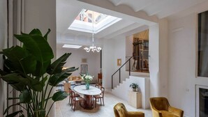 A spacious living area with a high ceiling, a skylight, a dining table with chairs, and a staircase leading to an upper level.