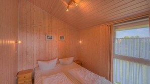 A wooden cabin bedroom with a bed, nightstands, and a window with curtains. - Ferienhaus in Rechlin (Rechlin)